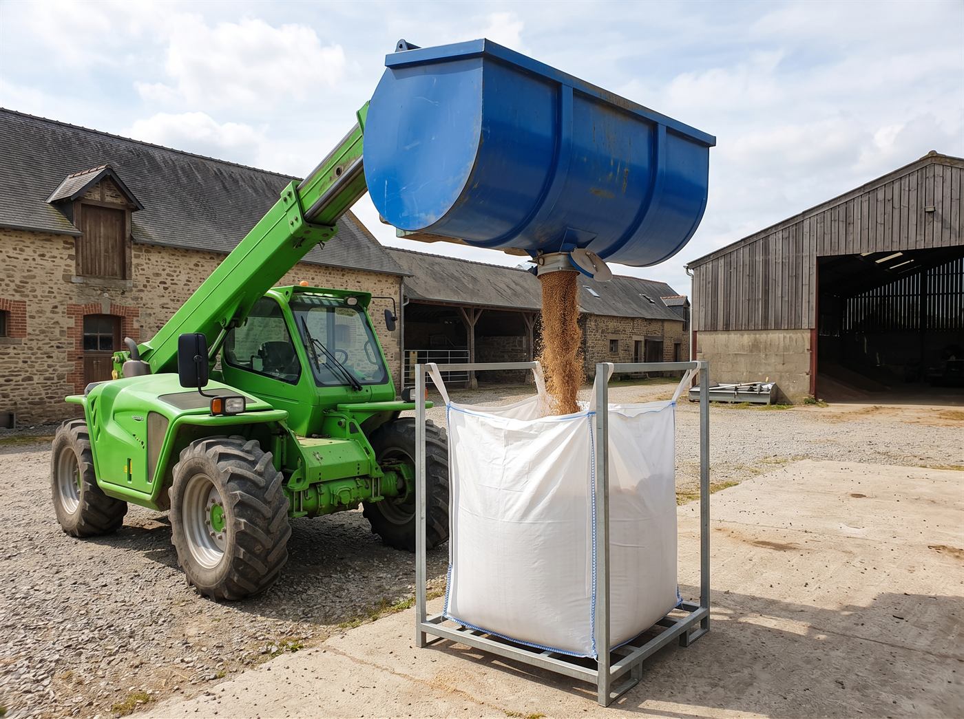 Un chargeur télescopique tient un godet au-dessus d'un grand sac souple ouvert, dans une cour de ferme ; des grains coulent de la vanne située sous le godet, droit dans le sac.