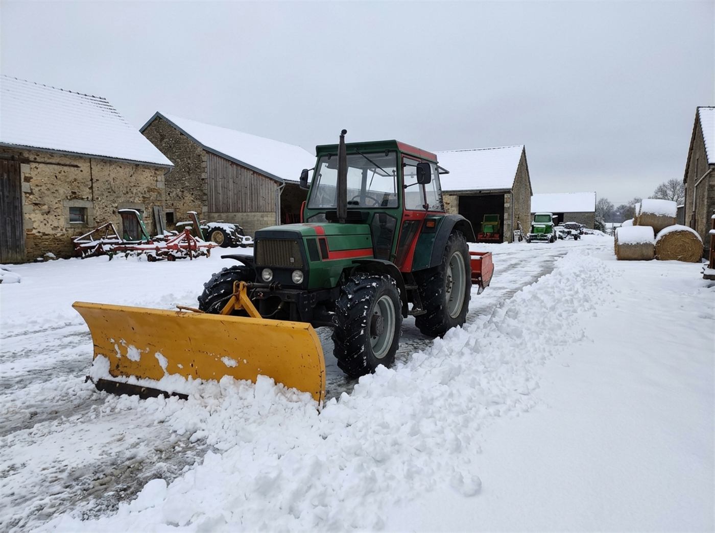 Un tracteur pousse une large lame de déneigement orientable sur une cour de ferme enneigée ; la neige est repoussée sur le côté.