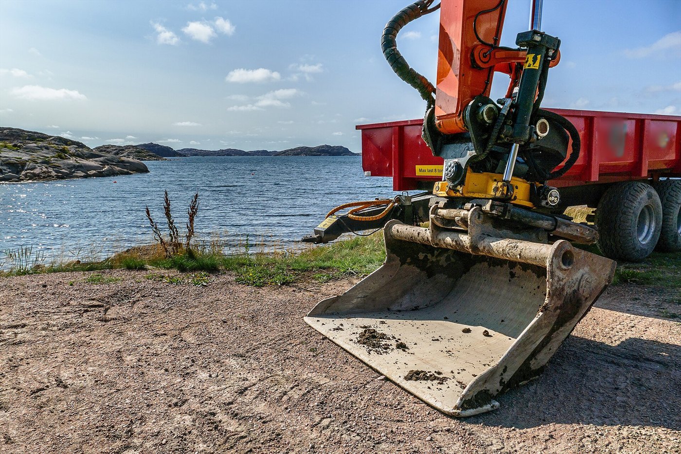 Godet de pelle posé sur une plage de gravier au bord de la mer, relié au balancier orange par une attache alimentée en flexibles ; une benne rouge à l'arrière-plan.