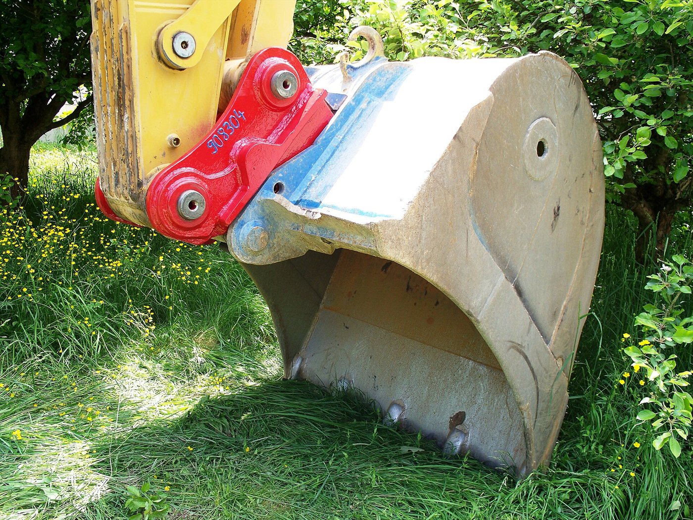 Godet beige suspendu au balancier jaune d'une pelle, relié par une attache rouge portant deux axes, devant de l'herbe et des arbustes.