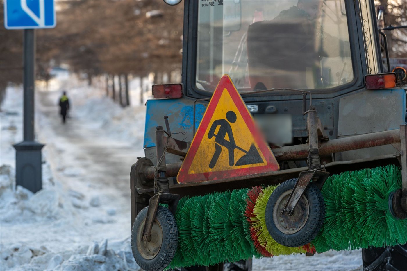 Avant d'un tracteur bleu portant une brosse rotative verte et jaune au ras du sol, sur un trottoir enneigé ; un panneau de travaux est fixé au-dessus de la brosse.