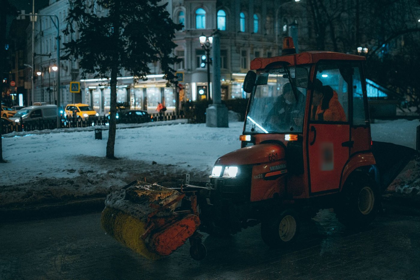 Petit tracteur orange avec cabine, brosse rotative à l'avant, sur une place enneigée de nuit, devant des façades éclairées.