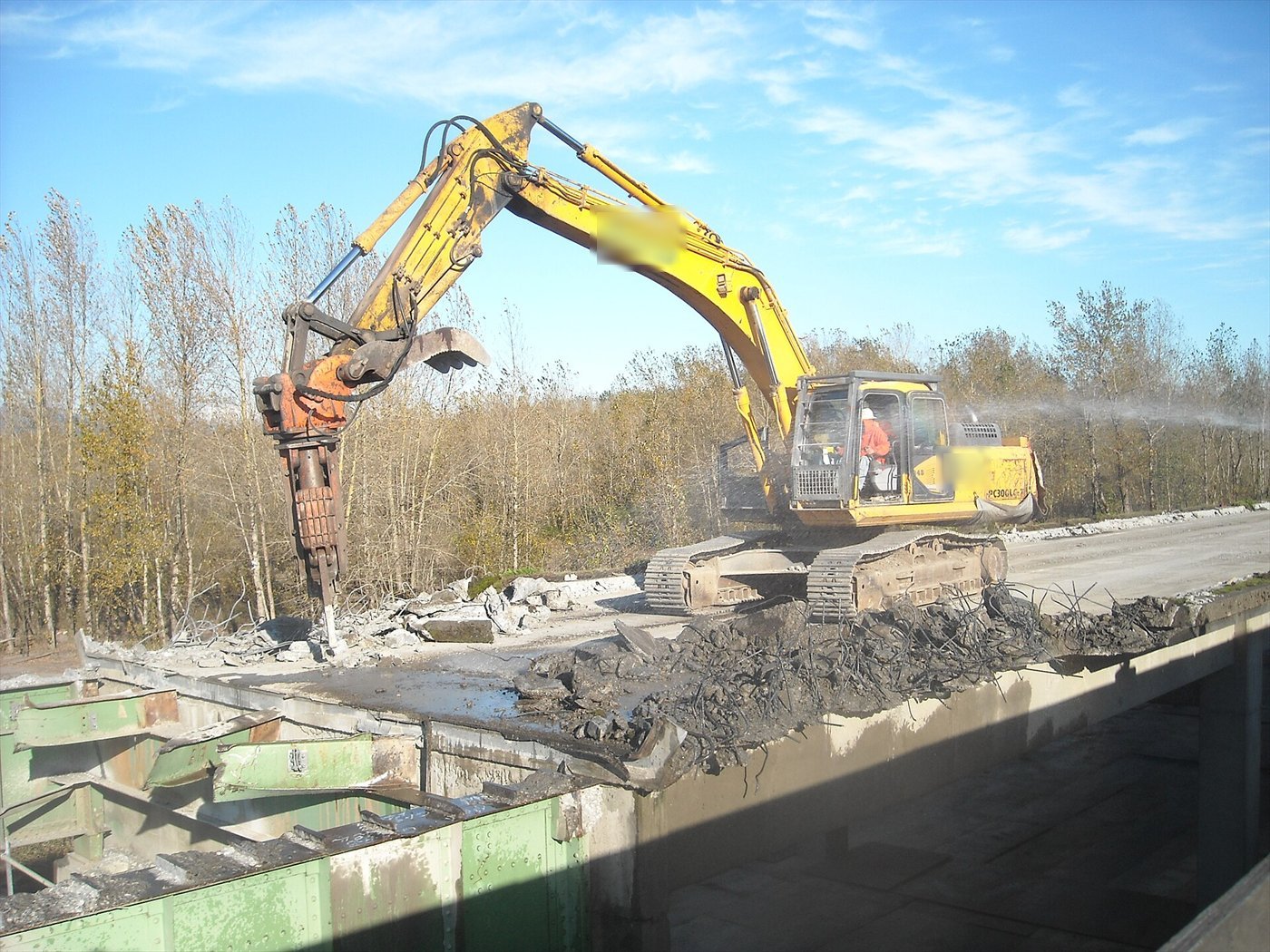 Grande pelle jaune sur le tablier d'un pont en démolition, son brise-roche orange enfoncé dans le béton ; un jet d'eau abat la poussière.