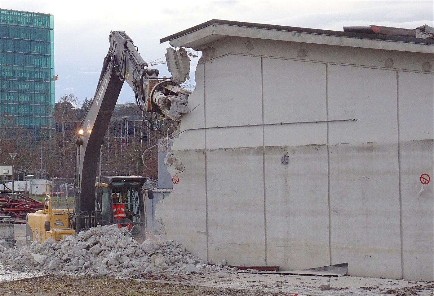 Pelle de démolition à long bras, croqueuse en tête, attaquant le haut d'un mur en béton, gravats au pied ; un immeuble vitré à l'arrière-plan.