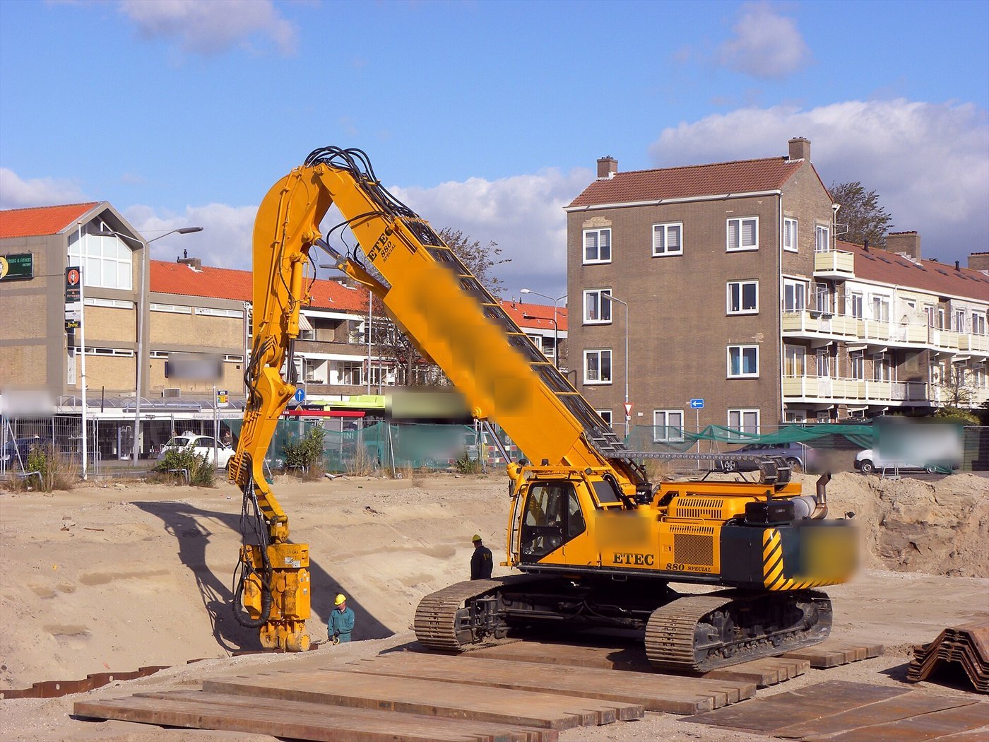 Pelle orange à grand bras, un vibrofonceur jaune au bout de la flèche tenant une palplanche ; des plaques au sol et des immeubles derrière.