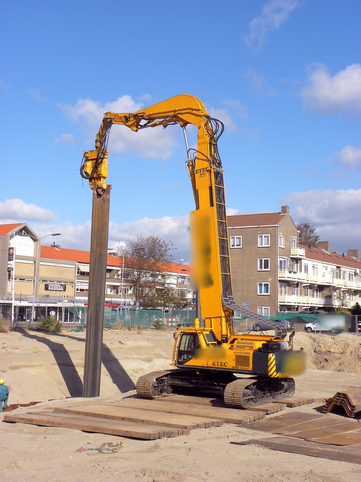Pelle orange à grand bras levant une longue palplanche à la verticale, tenue par un vibrofonceur, sur un chantier sablonneux en ville.