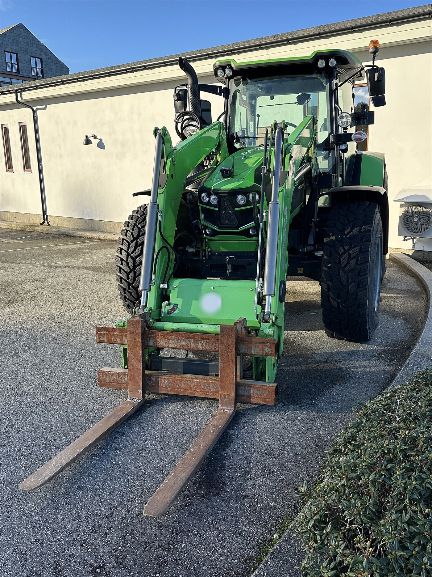 Tracteur vert vu de face, chargeur frontal abaissé portant un tablier et deux fourches à palettes, sur un sol goudronné.