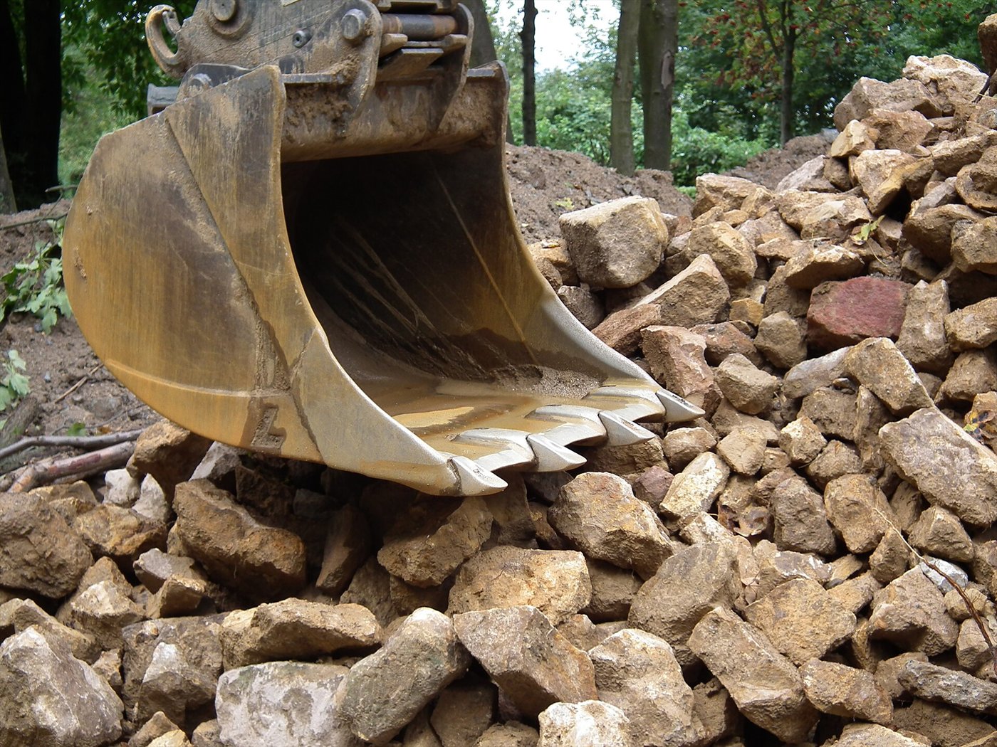Godet de pelle jaune, rangée de dents vers l'avant, posé sur un tas de blocs de grès, devant des arbres.