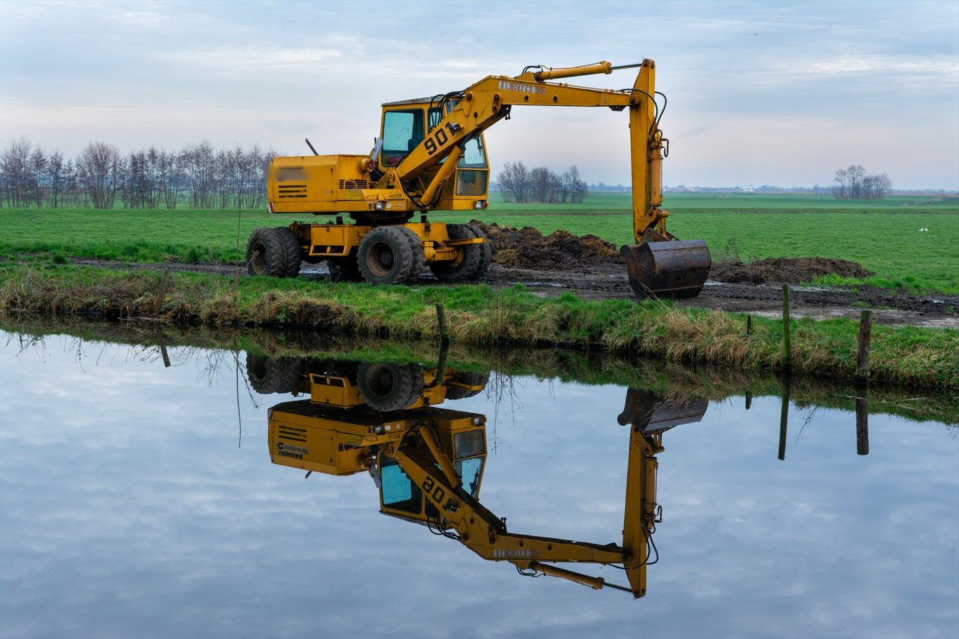 Pelle jaune sur pneus au bord d'un canal, bras tendu, un godet large posé sur la berge ; la machine se reflète dans l'eau.