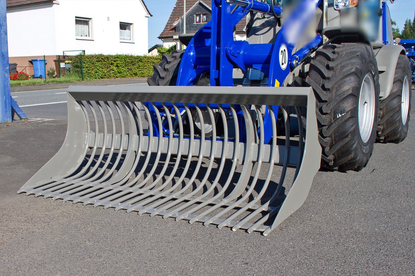 Grauer Gitterlöffel an einem kleinen blauen Lader, im Dreiviertelprofil, auf einer Dorfstraße.
