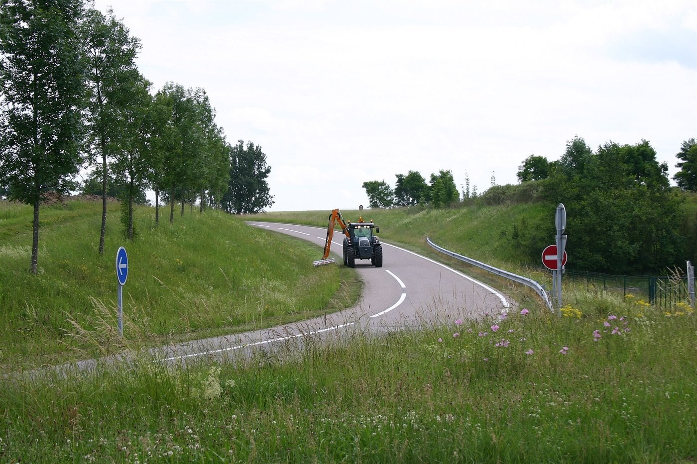 Tracteur avançant dans un virage de route de campagne, un bras articulé orange déporté sur la droite passe une tête de broyage sur l'accotement herbeux.