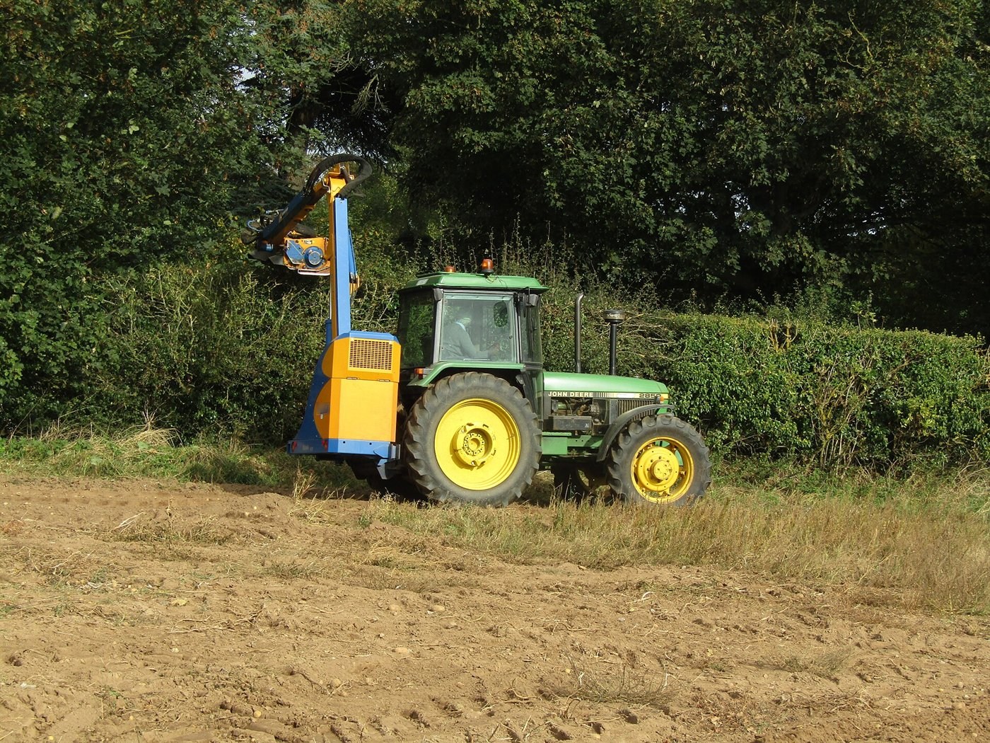 Tracteur vert au bord d'un champ labouré, un bras articulé jaune et bleu levé à la verticale taille une haie haute.