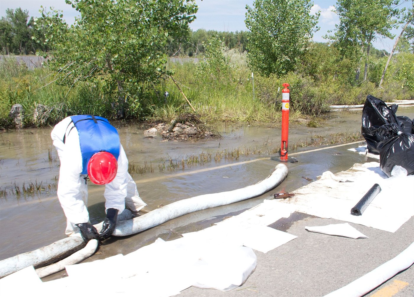 Intervenant en combinaison blanche et casque rouge, vu de dos, posant un long boudin absorbant au bord d'une eau trouble ; des feuilles absorbantes blanches couvrent la chaussée.