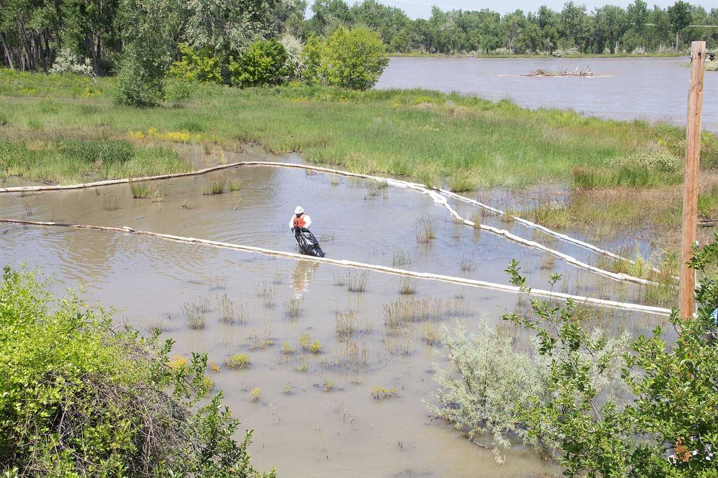 Zone humide inondée fermée par une ligne de boudins absorbants blancs ; un intervenant au loin remplit un sac noir.