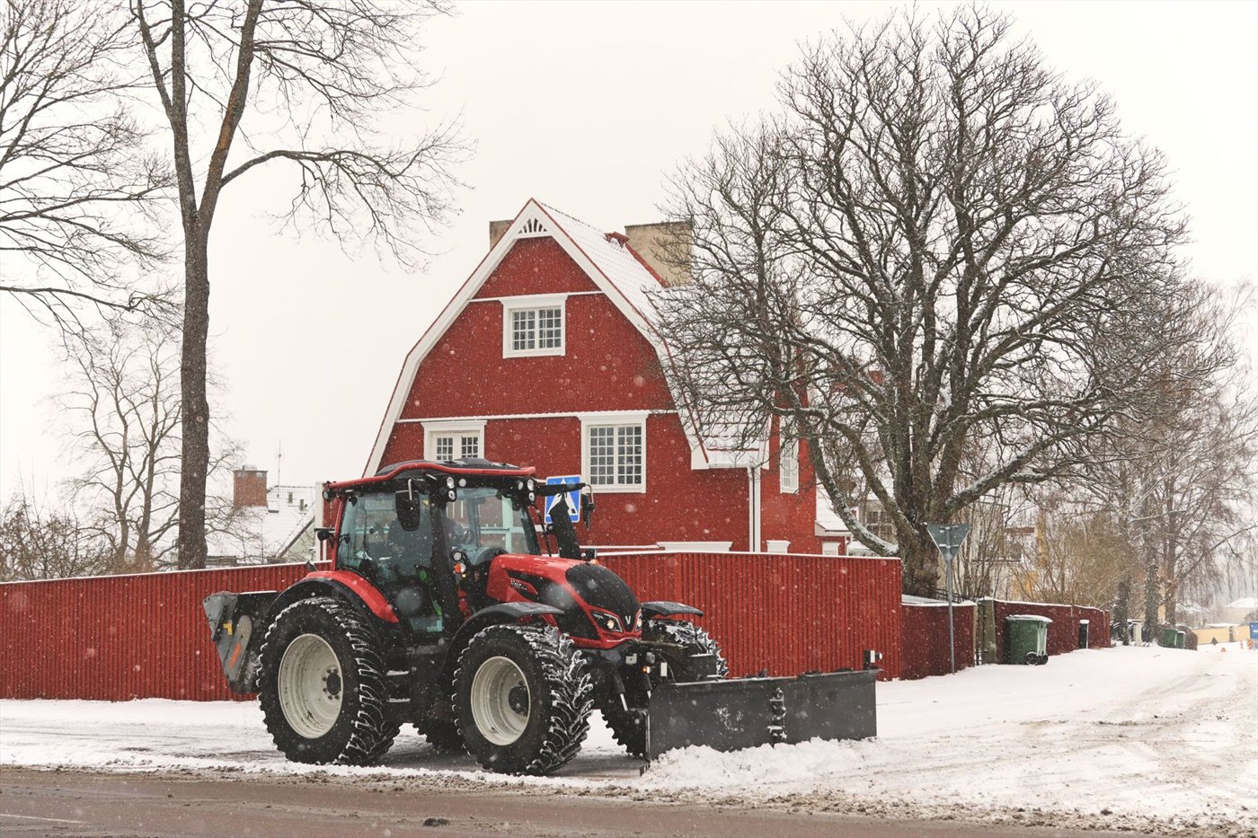 Tracteur rouge muni d'une lame de déneigement noire à l'avant, sur une route enneigée, devant une maison suédoise en bois rouge.