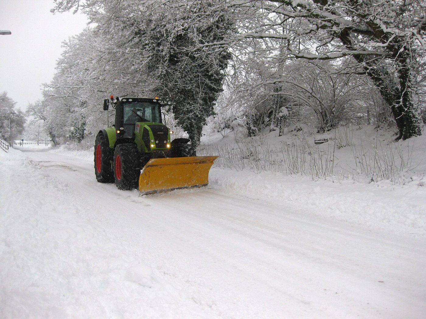 Tracteur vert avec une lame de déneigement jaune orientée en biais, sur une route de campagne enneigée, entre des arbres givrés.