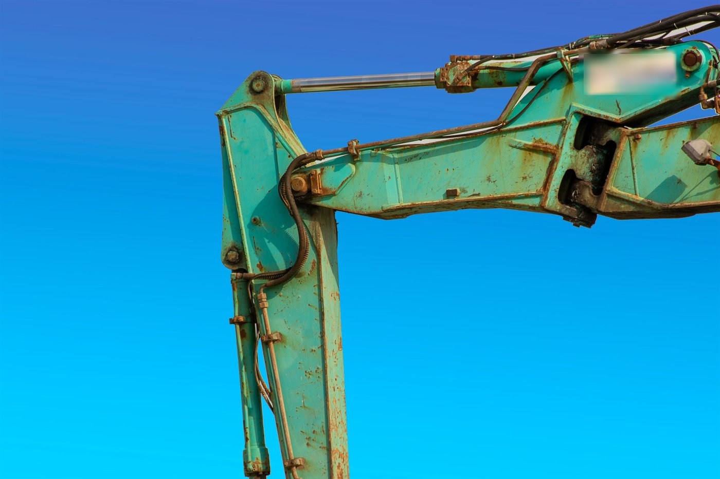 Bras de pelle usé vu de profil sur ciel bleu ; des flexibles hydrauliques courent le long de la flèche et du balancier, maintenus par des colliers.