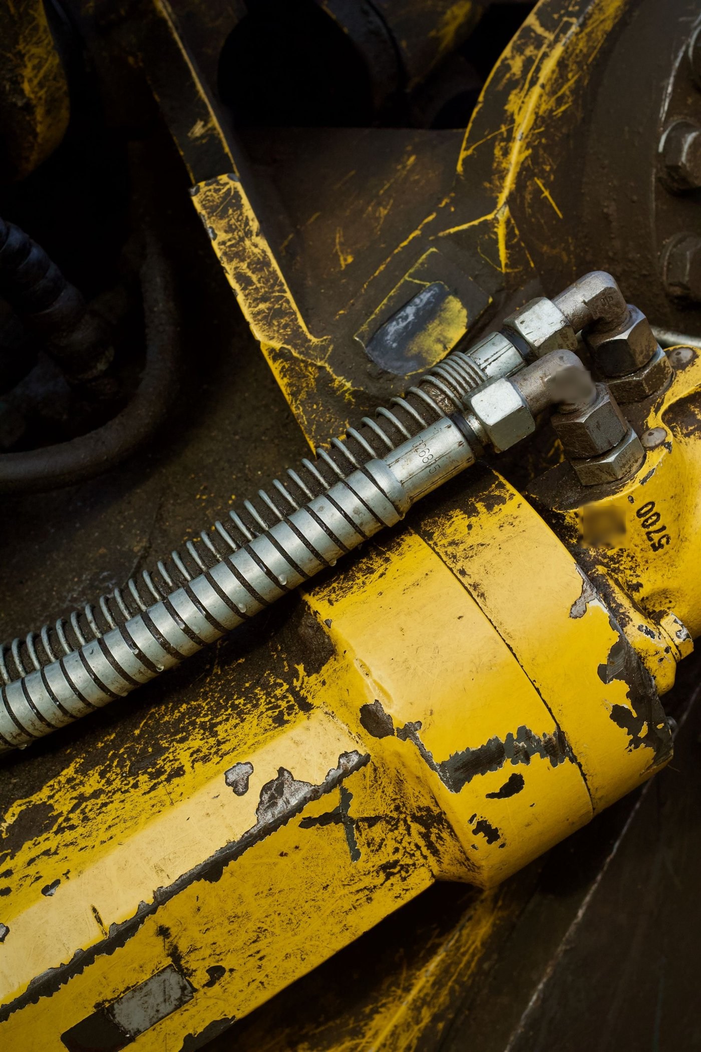 Gros plan de deux flexibles hydrauliques protégés par un ressort, raccordés par des coudes vissés sur un vérin jaune écaillé.