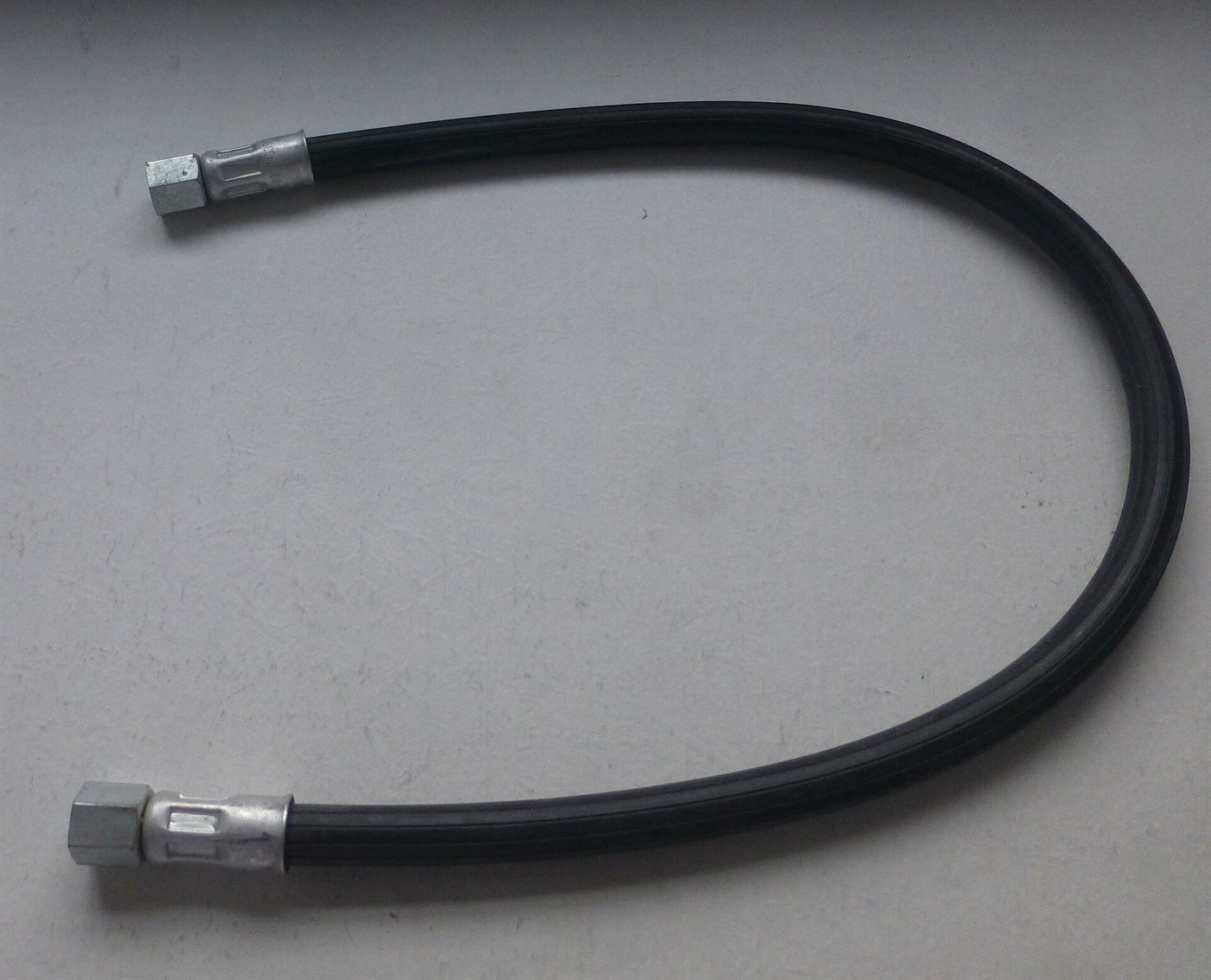Flexible hydraulique noir posé en arc sur un fond gris, avec un écrou et une bague sertie à chaque bout.