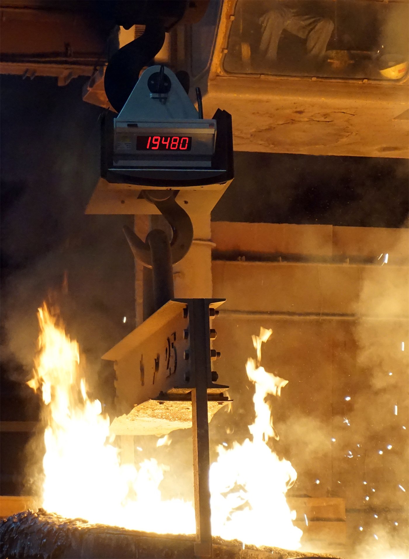Peson électronique suspendu à un crochet de pont roulant, son afficheur rouge allumé, une charge métallique accrochée dessous au-dessus de flammes de fonderie.