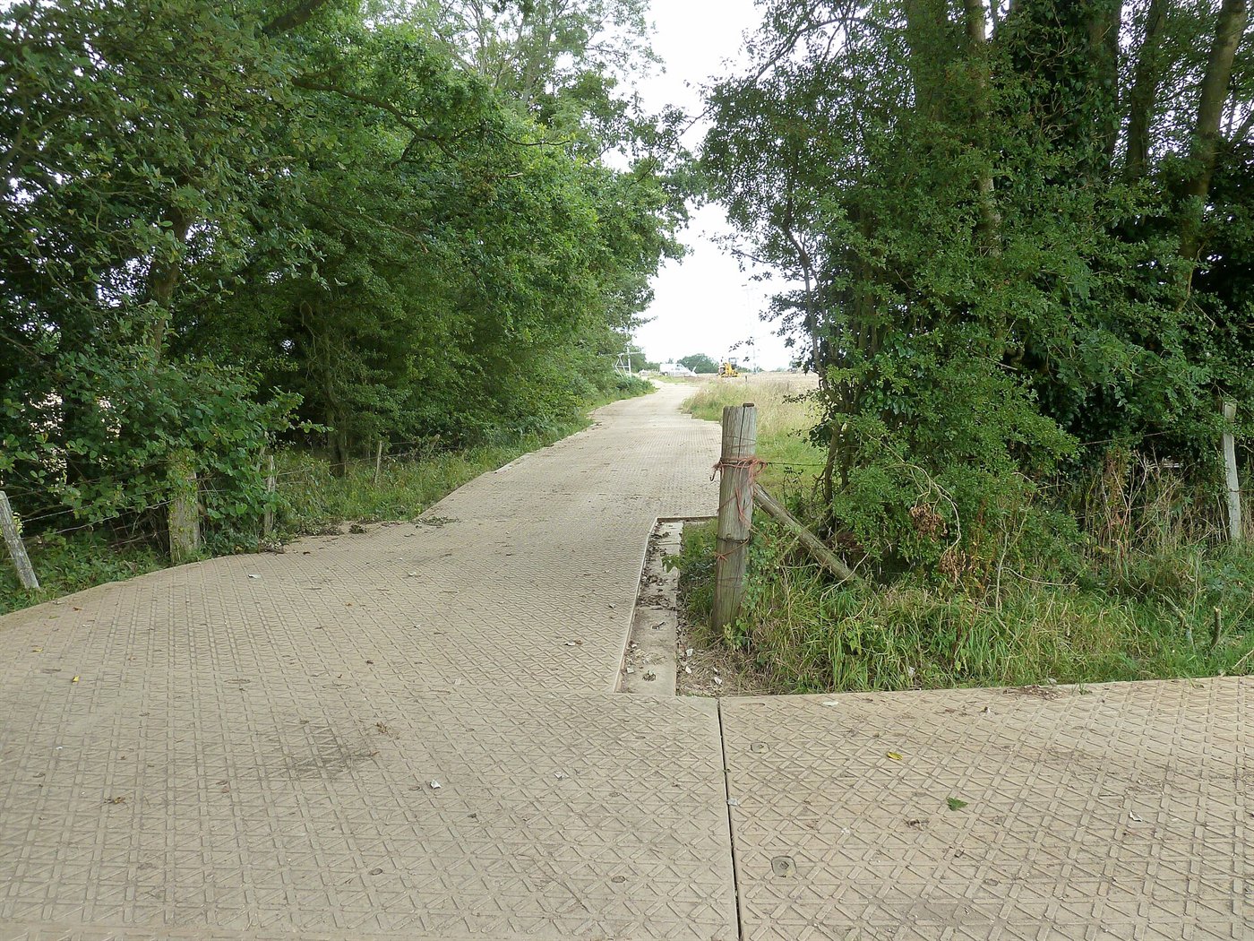 Piste provisoire faite de grandes plaques à relief antidérapant posées bord à bord entre deux haies, menant vers un chantier au loin.