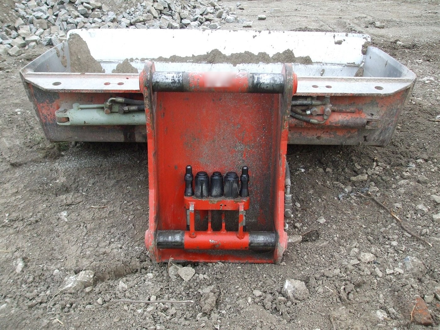 Red coupler plate fitted to a tilting bucket resting on the ground; between the two pins, a block carries five hydraulic couplings in a row.