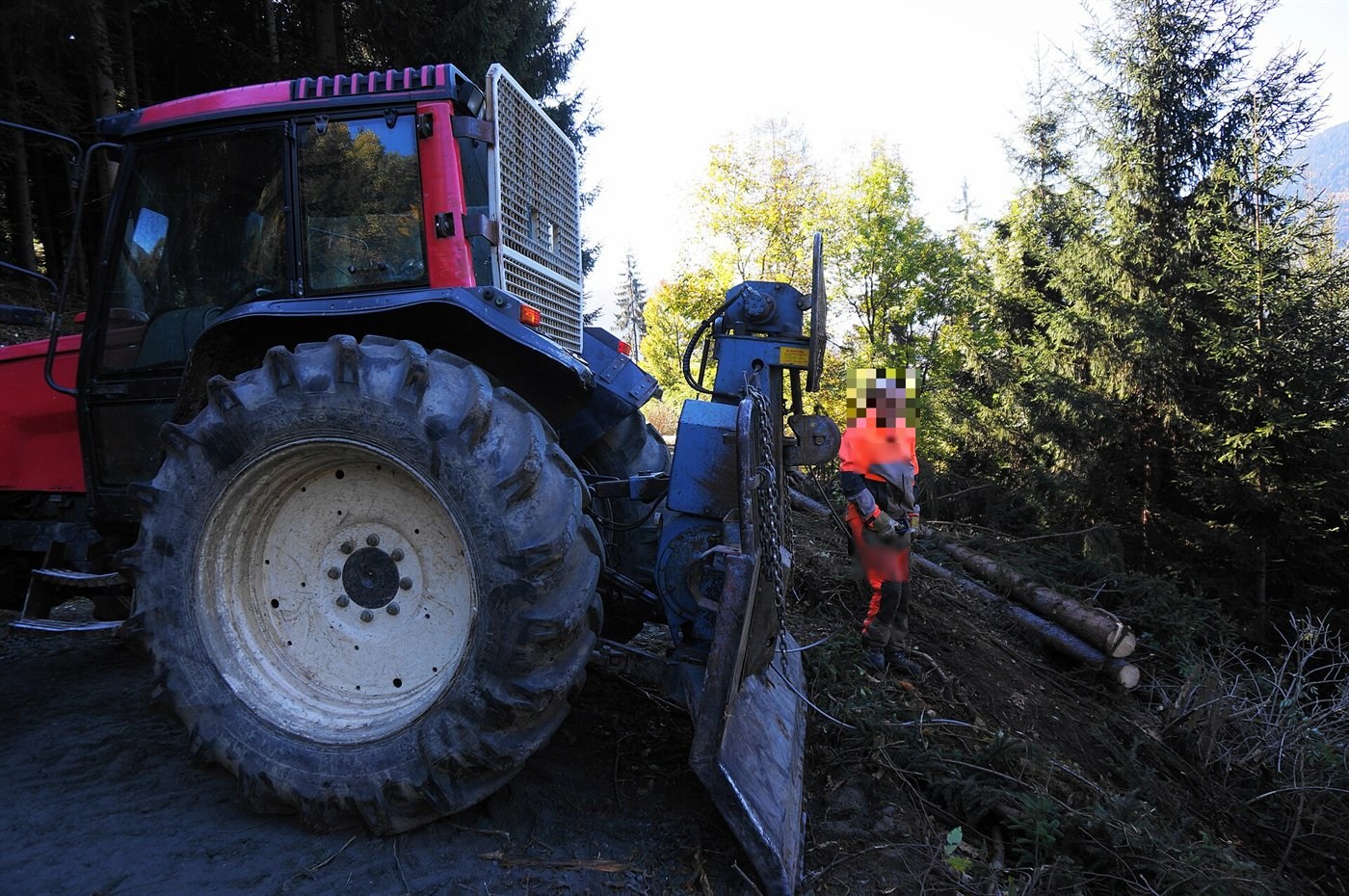 Tracteur forestier rouge vu de l'arrière, un treuil à lame d'appui posé au sol au bord de la piste ; un bûcheron en tenue orange se tient à côté, des grumes sur la pente.